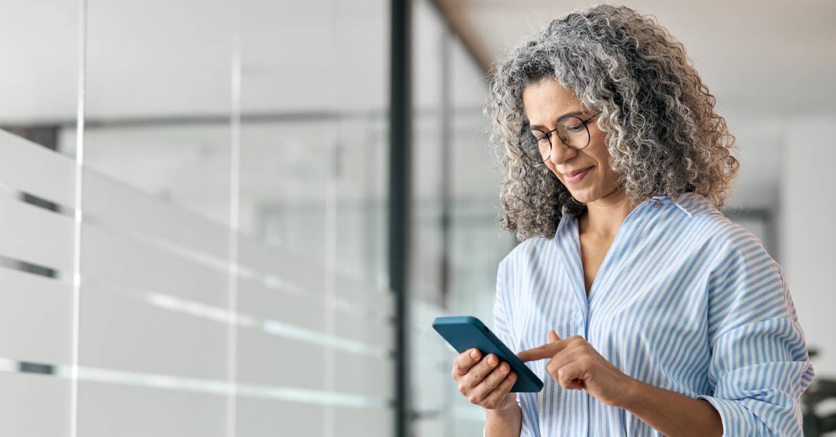 A middle-aged woman wearing a striped button-down blouse looks down and scrolls through the banking app on her phone.