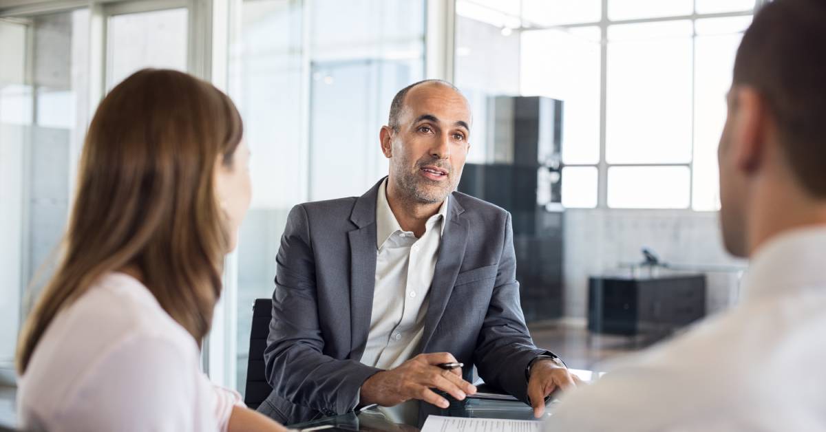A financial advisor in a suit sitting at an office desk, advising a couple sitting across from him.